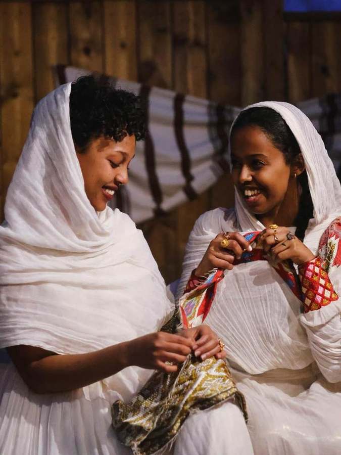 Two women in white shawls smiling, holding woven cloth