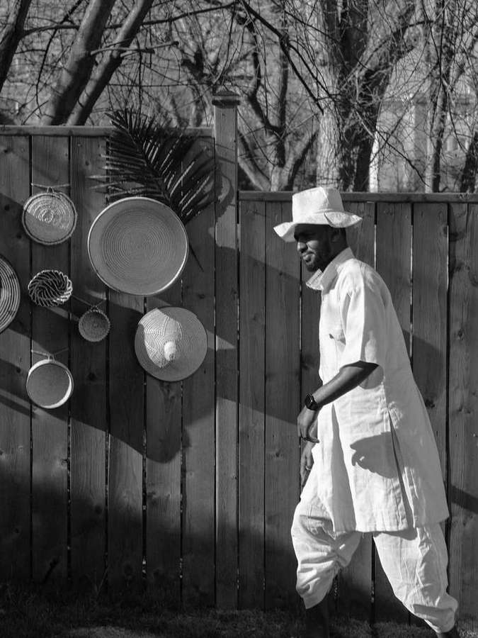 A man in a white hat and long shirt walking past a fence of hanging baskets
