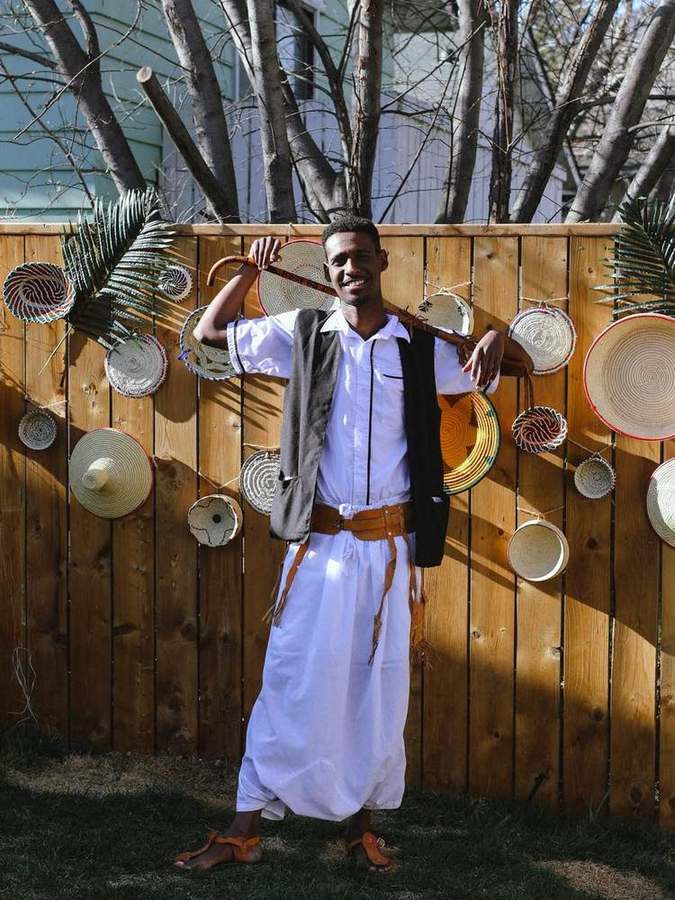 A man in a white shirt and wrapped skirt holding a cane, standing by a decorated fence